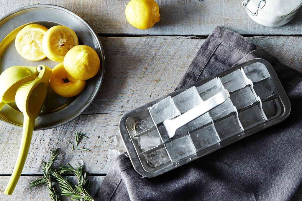 A stainless steel ice cube tray next to a bowl of squeezed lemons